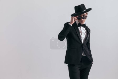Téléchargez les photos : Élégant jeune marié en smoking noir portant un chapeau et des lunettes de soleil ajustables, debout et posant la main dans la poche devant un fond gris - en image libre de droit
