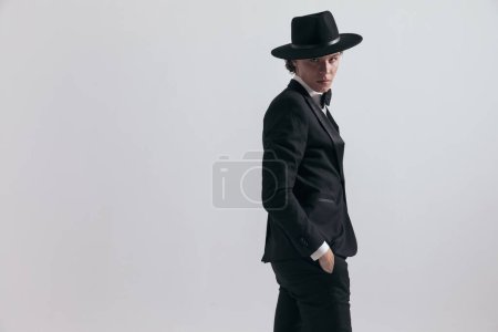 Photo for Portrait of elegant best man wearing hat and posing with hands in pockets in front of grey background in studio, side view - Royalty Free Image
