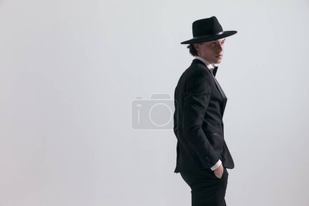 Photo for Side view of handsome young groom in black tuxedo with hat looking to side and posing with hands in pockets in front of grey background - Royalty Free Image