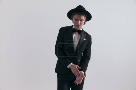 Téléchargez les photos : Élégant homme sexy en smoking noir avec chapeau toucher et frotter les paumes en face de fond gris en studio - en image libre de droit