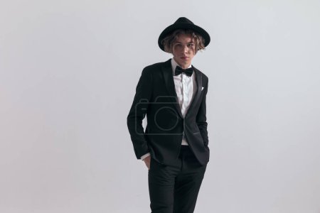Photo for Elegant groom wearing black tuxedo and posing with hands in pockets in front of grey background in studio - Royalty Free Image