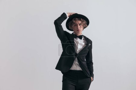 Téléchargez les photos : Jeune homme élégant aux cheveux longs en smoking noir ajustant le chapeau et posant la main dans la poche devant fond gris en studio - en image libre de droit