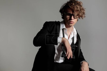 Téléchargez les photos : Sexy gars de la mode en smoking avec chemise à col ouvert avec des lunettes tenant les bras sur le genou en face de fond gris en studio - en image libre de droit