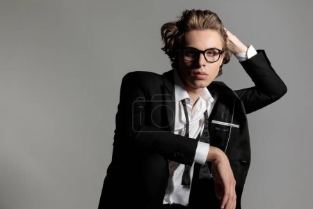 Téléchargez les photos : Élégant jeune homme avec des lunettes ajustant les cheveux longs et posant d'une manière fraîche en face de fond gris - en image libre de droit