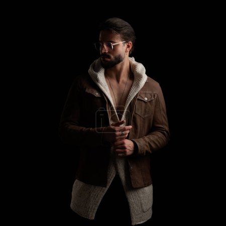 Téléchargez les photos : Mode jeune homme ne portant que cardigan de laine et veste et regardant de côté tout en posant d'une manière fraîche en face de fond noir - en image libre de droit