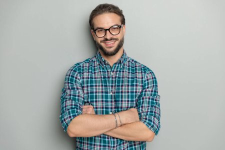Foto de Retrato de hombre nerd feliz con gafas con camisa a cuadros, cruzando brazos y sonriendo mientras posando frente a fondo gris en el estudio - Imagen libre de derechos