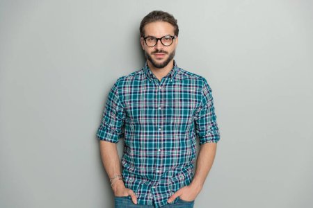 Foto de Retrato de chico sexy casual con camisa a cuadros y gafas y posando con las manos en los bolsillos delante de fondo gris en el estudio - Imagen libre de derechos