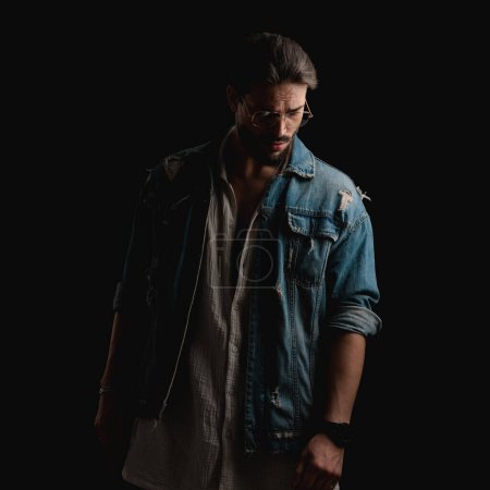Photo for Fashion casual man wearing glasses and denim jacket looking down and posing in front of black background in studio - Royalty Free Image