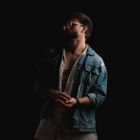 Photo for Portrait of sexy bearded man with opened shirt in denim jacket looking to side and posing in a cool way on black background - Royalty Free Image
