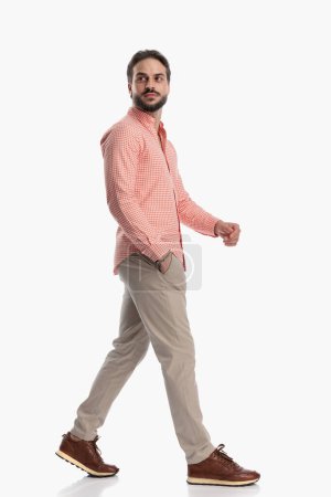 Photo for Side view of attractive bearded man looking away and walking with hand in pocket in front of white background - Royalty Free Image