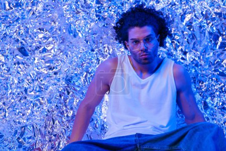 Photo for Fashion man in white t-shirt with glasses posing in a cool way while sitting in front of silver tinfoil background with blue light - Royalty Free Image