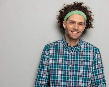 Téléchargez les photos : Portrait de gars heureux occasionnel avec des lunettes et bandeau souriant et posant tout en portant bandeau de sport sur fond gris - en image libre de droit