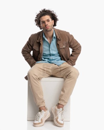 Téléchargez les photos : Confiant bouclé gars cheveux en veste beige tenant les mains dans les poches et posant d'une manière cool tout en étant assis sur fond blanc - en image libre de droit