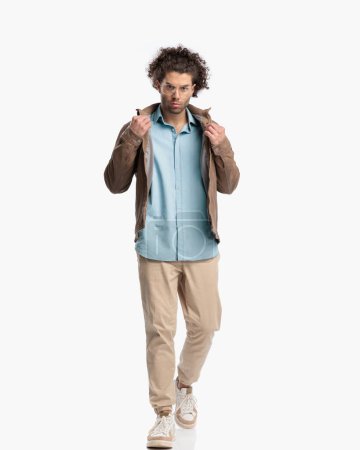 Photo for Full body picture of cool casual guy walking and adjusting jacket in front of white background in studio - Royalty Free Image