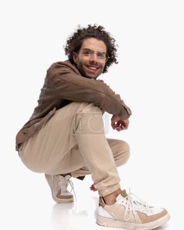 Photo for Happy young man with glasses and curly hair crouching with elbow on knee in front of white background - Royalty Free Image