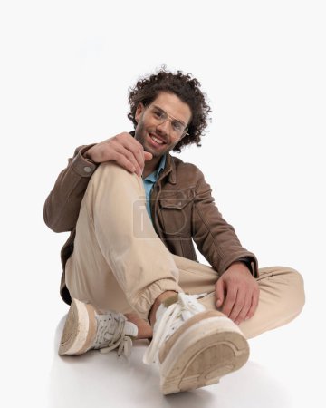 Photo for Happy casual guy with glasses and curly hair touching knee and sitting in front of white background - Royalty Free Image