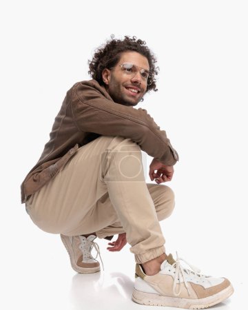 Téléchargez les photos : Vue latérale de jeune homme sexy avec des lunettes regardant loin et accroupi avec le coude sur le genou et souriant devant fond blanc - en image libre de droit