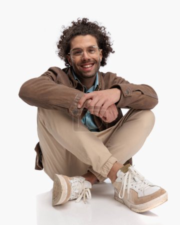 Photo for Excited casual man with glasses and curly hair laughing and holding hands while elbows are on knees in front of white background - Royalty Free Image