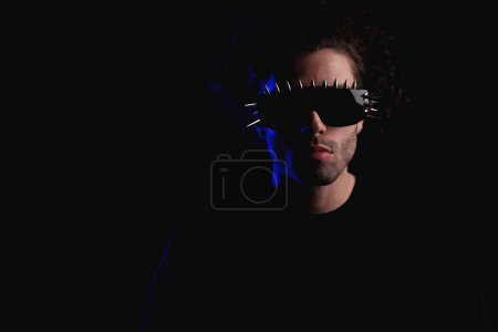 Téléchargez les photos : Portrait de jeune homme décontracté aux cheveux bouclés portant des lunettes de soleil fraîches et posant devant un fond noir - en image libre de droit