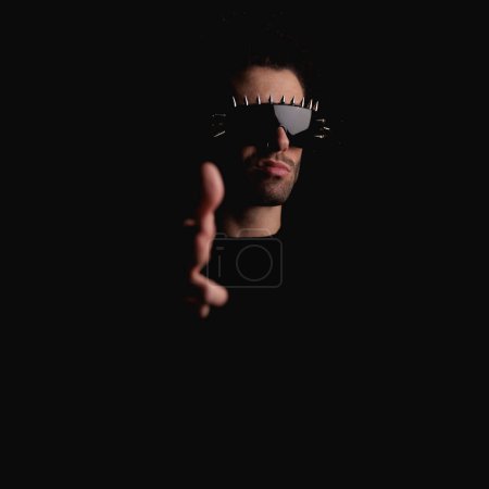 Photo for Fashion male model wearing cool sunglasses and pointing hand forward posing in a confident way in front of black background - Royalty Free Image