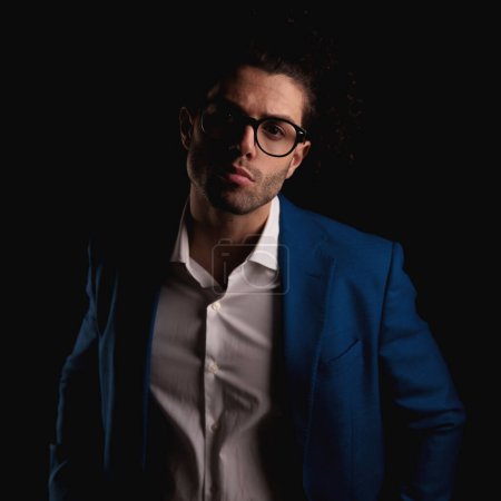 Téléchargez les photos : Portrait d'un jeune homme séduisant avec chemise à col ouvert portant un costume bleu et des lunettes posant de manière confiante devant un fond noir - en image libre de droit