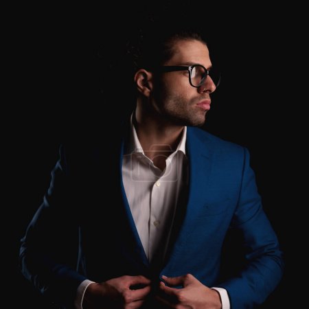 Téléchargez les photos : Homme élégant confiant avec des lunettes et col ouvert chemise boutonnage costume tout en regardant de côté en face de fond noir - en image libre de droit
