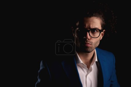 Téléchargez les photos : Bouleversé jeune homme avec des cheveux bouclés et des lunettes portant costume et fronçant les sourcils tout en regardant vers l'avant en face de fond noir en studio - en image libre de droit