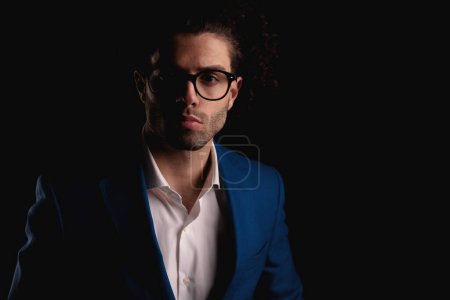 Téléchargez les photos : Portrait d'un bel homme d'affaires avec des lunettes en costume bleu et des lunettes avec chemise à col ouvert posant devant un fond noir - en image libre de droit