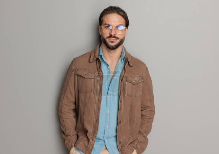 Foto de Retrato de hombre casual con chaqueta de cuero marrón y camisa de mezclilla, tomados de la mano en bolsillos y posando delante de fondo gris claro - Imagen libre de derechos