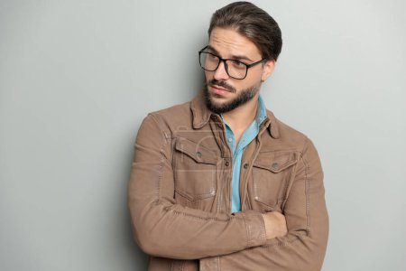 Photo for Portrait of handsome young man with glasses wearing brown leather jacket folding arms and looking to side in front of grey background in studio - Royalty Free Image