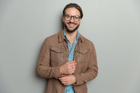 Téléchargez les photos : Homme décontracté heureux avec des lunettes portant une veste en cuir marron et ajustant les manches, souriant et posant devant un fond gris - en image libre de droit