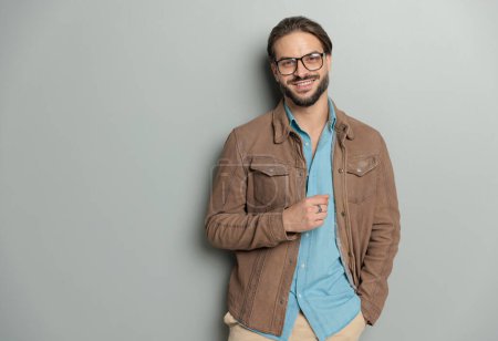 Téléchargez les photos : Portrait d'un homme décontracté heureux tenant la main dans la poche et souriant tout en portant une veste en cuir marron et une chemise en denim devant un fond gris - en image libre de droit