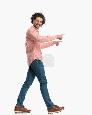 Téléchargez les photos : Vue latérale de heureux cheveux bouclés homme souriant et pointant du doigt tout en marchant devant fond blanc en studio - en image libre de droit