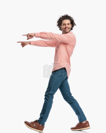 Photo for Side view of enthusiastic young man with curly hair pointing fingers to side and walking while being happy in front of white background in studio - Royalty Free Image