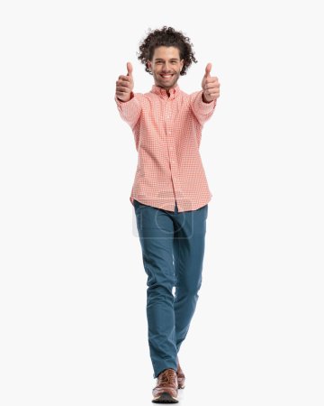 Téléchargez les photos : Homme heureux avec les cheveux bouclés souriant et faire des pouces geste tout en marchant devant fond blanc - en image libre de droit