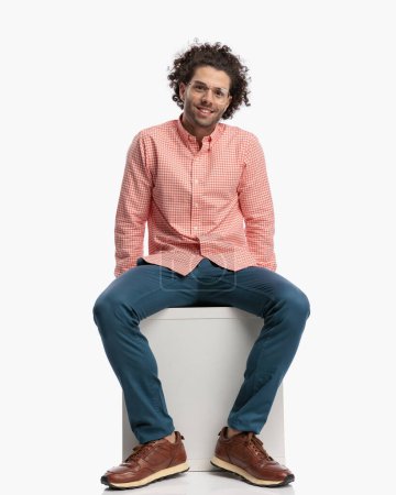 Téléchargez les photos : Image corporelle complète du jeune homme heureux avec des lunettes souriant et posant tout en étant assis devant fond blanc en studio - en image libre de droit