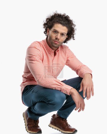 Photo for Sexy gashion man with glasses and curly hair crouching and holding arms on knees in front of white background - Royalty Free Image