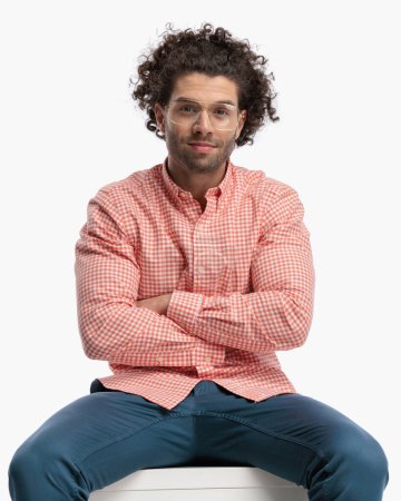 Photo for Proud casual man with curly hair and glasses smiling and folding arms while sitting in front of white background in studio - Royalty Free Image