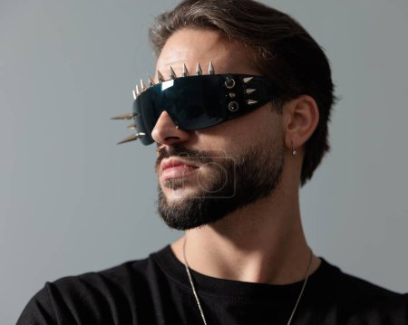 Téléchargez les photos : Portrait d'homme de mode barbu avec des lunettes de soleil fraîches regardant vers le côté et posant d'une manière fraîche sur fond gris - en image libre de droit