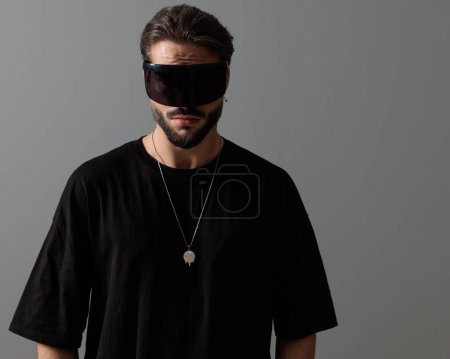 Téléchargez les photos : Cool homme de mode en t-shirt noir portant des lunettes et posant de manière confiante devant fond gris - en image libre de droit