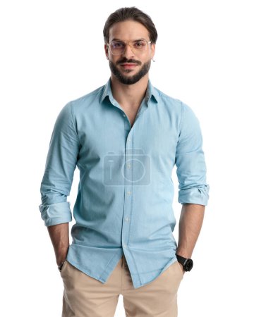 Foto de Atractivo hombre casual con camisa azul vaqueros cogidos de la mano en los bolsillos, mirando hacia adelante y sonriendo sobre fondo blanco - Imagen libre de derechos