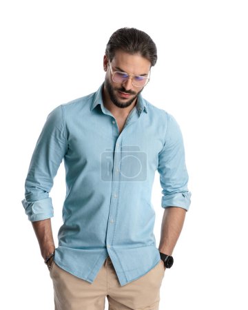 Foto de Sexy barbudo hombre con gafas cogidas de la mano en los bolsillos y mirando hacia abajo mientras posando sobre fondo blanco - Imagen libre de derechos