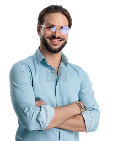 Foto de Atractivo joven con barba y gafas cruzando brazos y sonriendo delante de fondo blanco - Imagen libre de derechos