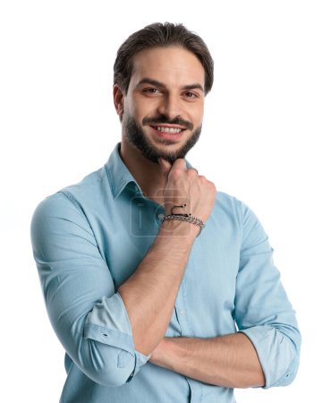 Foto de Feliz joven brazos plegables, sonriendo mientras toca la barba y posando delante de fondo blanco - Imagen libre de derechos