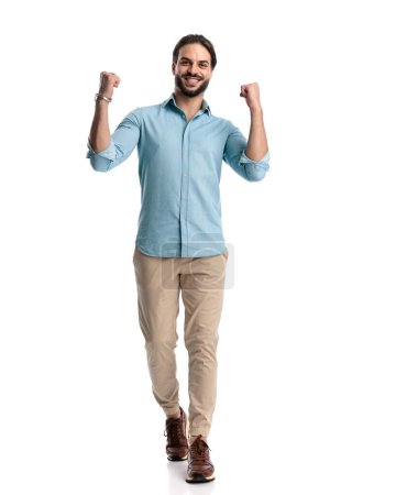 Photo for Excited casual man with beard holding fists up and celebrating victory, walking in front of white background in studio - Royalty Free Image