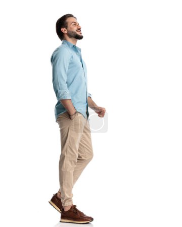 Téléchargez les photos : Vue latérale du gars heureux occasionnel marchant avec la main dans la poche et souriant devant fond blanc - en image libre de droit
