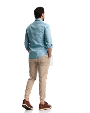 Photo for Behind view of casual man in denim shirt walking with hands in pockets in front of white background - Royalty Free Image