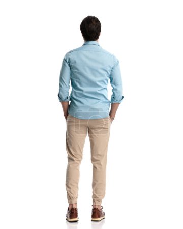 rear view of casual man in denim shirt with chino pants holding hands in pockets in front of white background 
