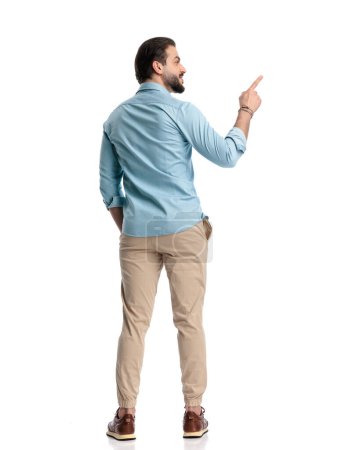 Foto de Atractivo hombre barbudo con la mano en el bolsillo apuntando dedo a lado y de pie delante de fondo blanco - Imagen libre de derechos