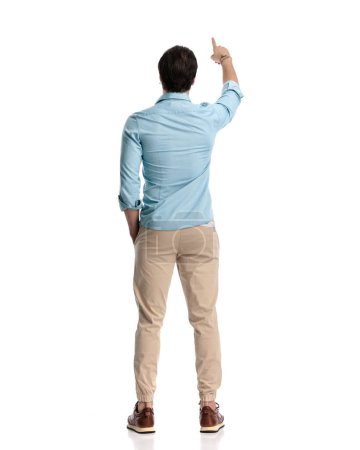 Photo for Back view of young man in denim shirt pointing finger and holding hands in pockets standing in front of white background - Royalty Free Image